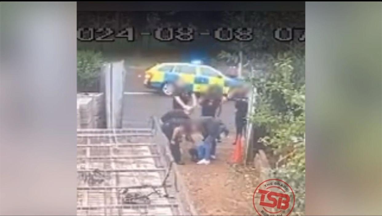 Five police officers bend over a man wearing a blue jacket who is face down on the ground. There is a wooden fence and hedge to the right and a police car is parked on the road behind