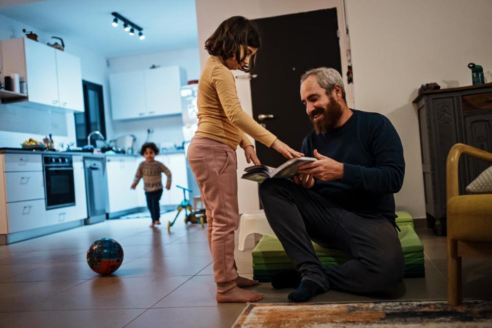 A bearded man holds an open book while seated and facing a girl who is standing. Another child plays in the background.