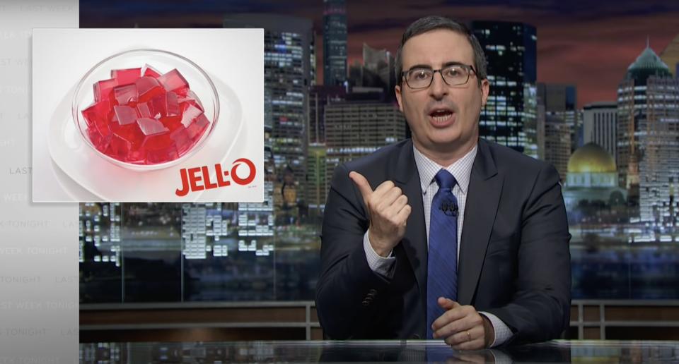 John Oliver at his desk, pointing to an image of Jello next to him