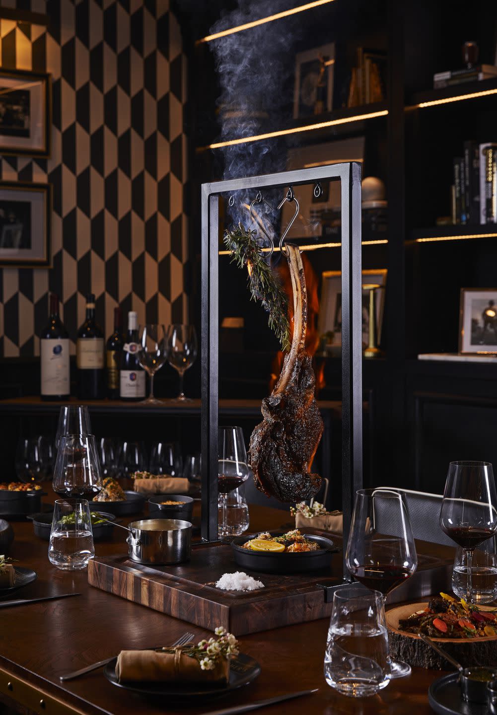 a tomahawk steak hanging from a device over a table with wine glasses and side dishes on it