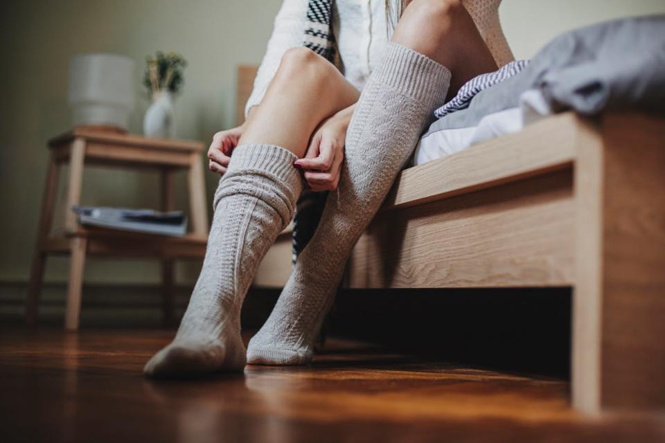 A woman sits on the edge of her bed to pull on a tall pair of wool socks