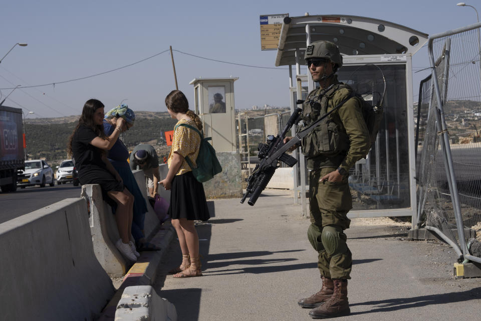 An Israeli soldier secures a bus stop while Israeli settlers wait for a ride at the Gush Etzion junction, the transportation hub for a number of West Bank Jewish settlements, Thursday, June 9, 2022. Israeli settlers in the occupied West Bank may soon have a taste of the military rule that Palestinians have been living under for 55 years. A looming end-of-month deadline to extend legal protections to Jewish settlers has put Israel’s government on the brink of collapse and drawn widespread warnings that the territory could be plunged into chaos. (AP Photo/Maya Alleruzzo)