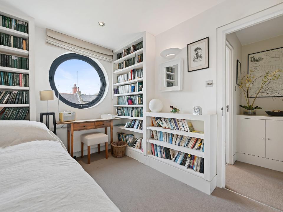 A white bedroom with the bed of the left, bookshelves on the left and right, and a circular window in the middle