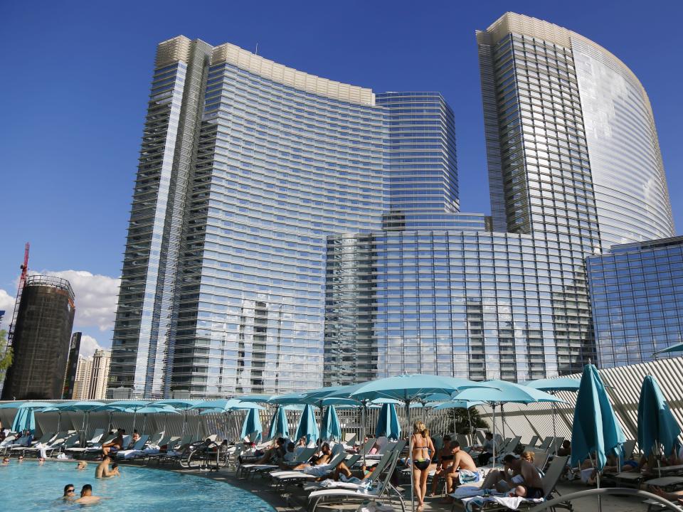 A hotel pool lined with loungers and blue umbrellas with skyscrapers behind.