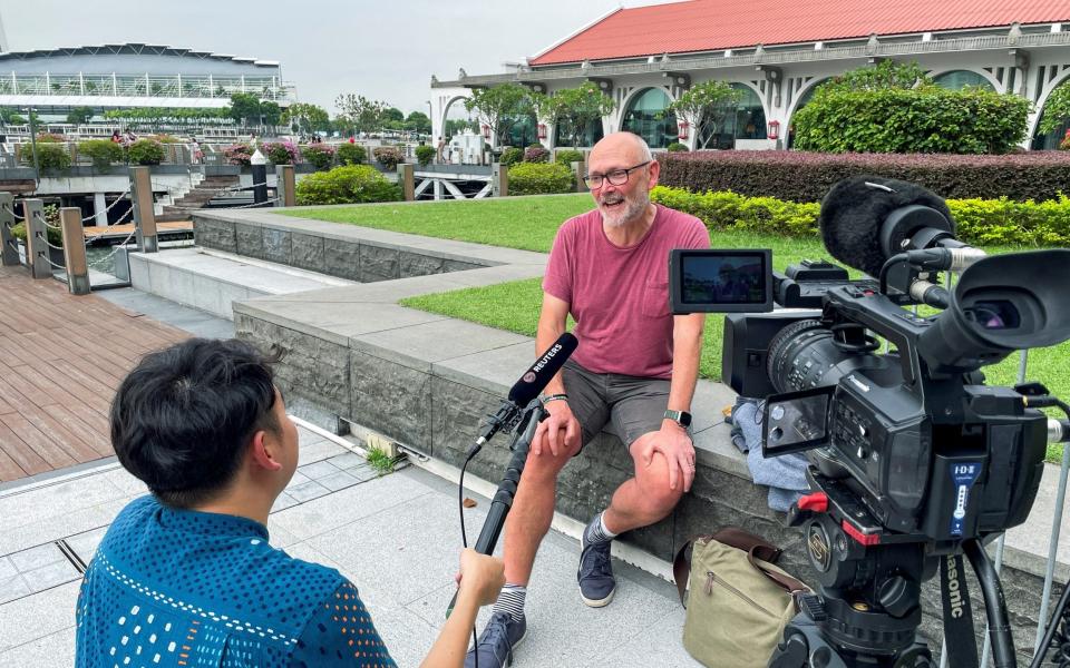 A man sits on a low wall in shorts being filmed and interviewed by a man holding a microphone marked 'Reuters'