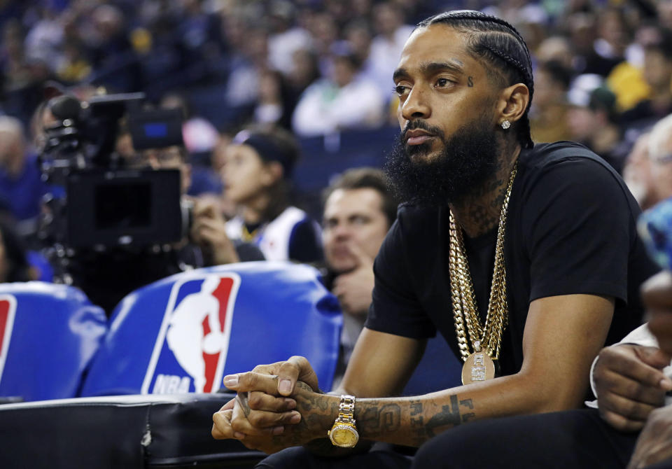 FILE - In this March 29, 2018, file photo, rapper Nipsey Hussle watches an NBA basketball game between the Golden State Warriors and the Milwaukee Bucks in Oakland, Calif. Hussle, 33, was shot and killed outside his Los Angeles clothing store on March 31, 2019. A year after Hussle's death, his popularity and influence are as strong as ever. He won two posthumous Grammys in January, he remains a favorite of his hip-hop peers and his death has reshaped his hometown of Los Angeles in some unexpected ways. (AP Photo/Marcio Jose Sanchez, File)