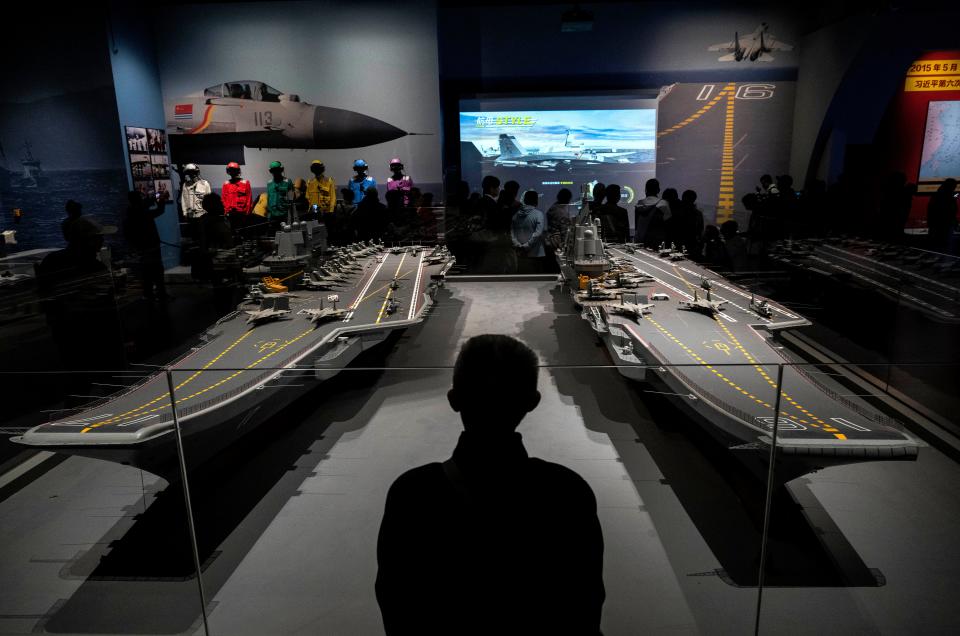 A man stands in front of two models of Chinese aircraft carriers, depicting model fighter jets, in a museum with other exhibits and displays highlighting Chinese naval aviation.