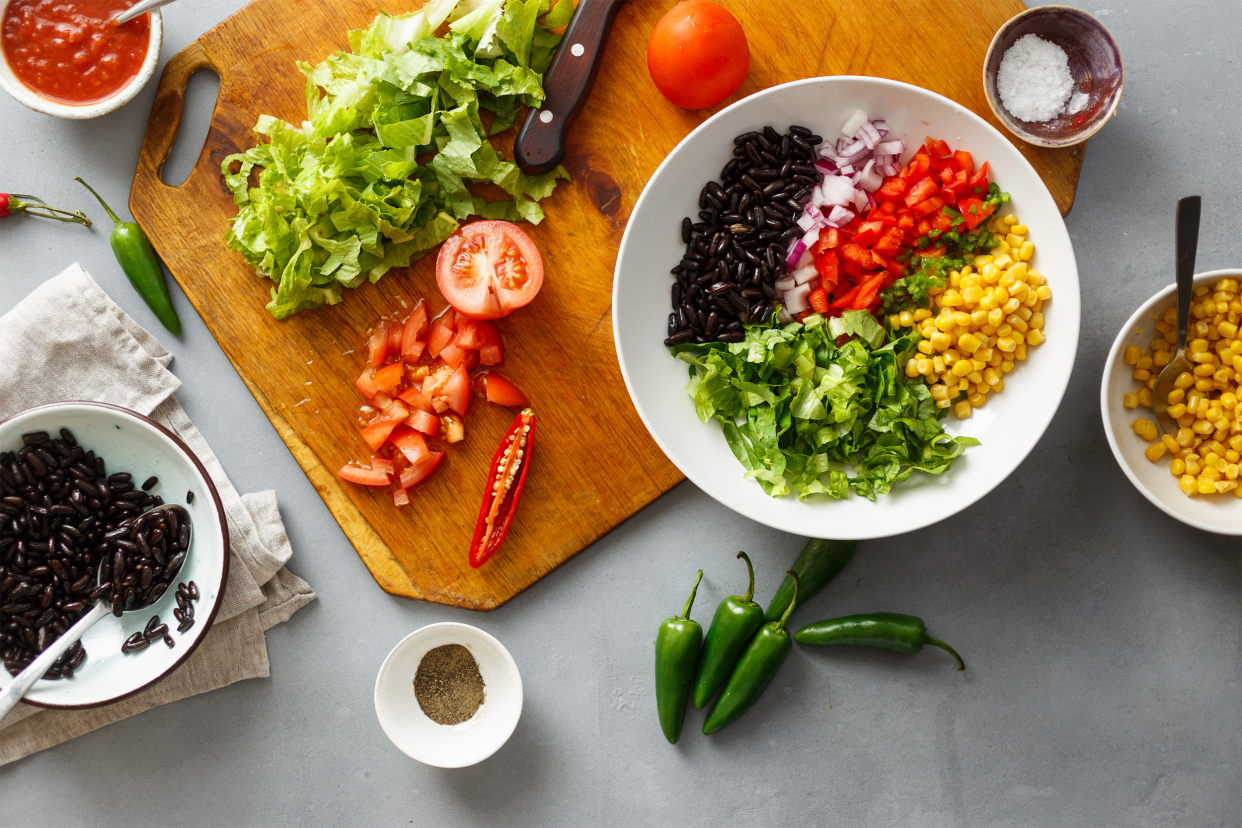 Top-view of several different ingredients for meatless taco salad, in bowls, a plate, and a wooden cutting board on a grey stone table