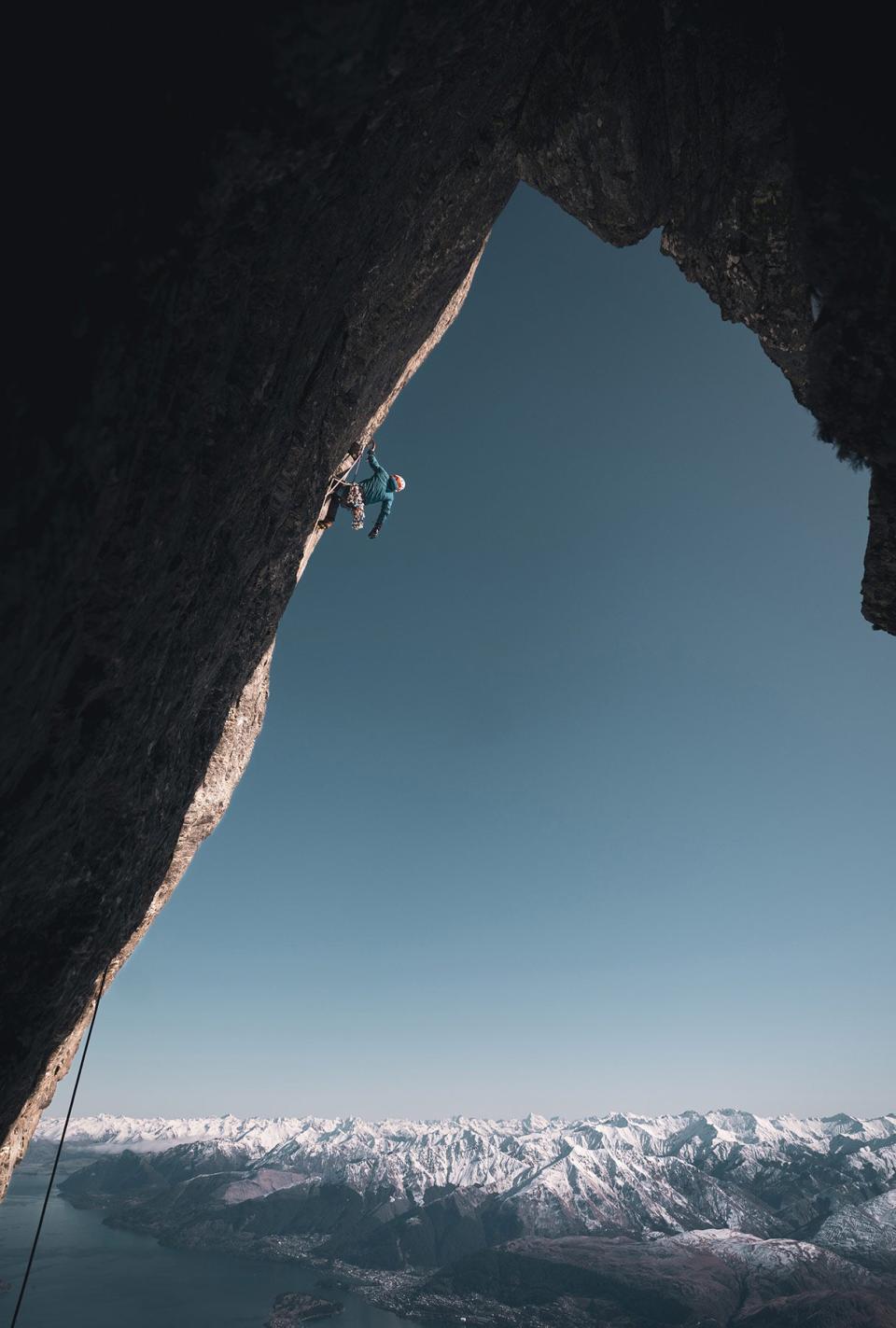 A mountain climber scales a steep cliff with snow-covered mountains in the background.