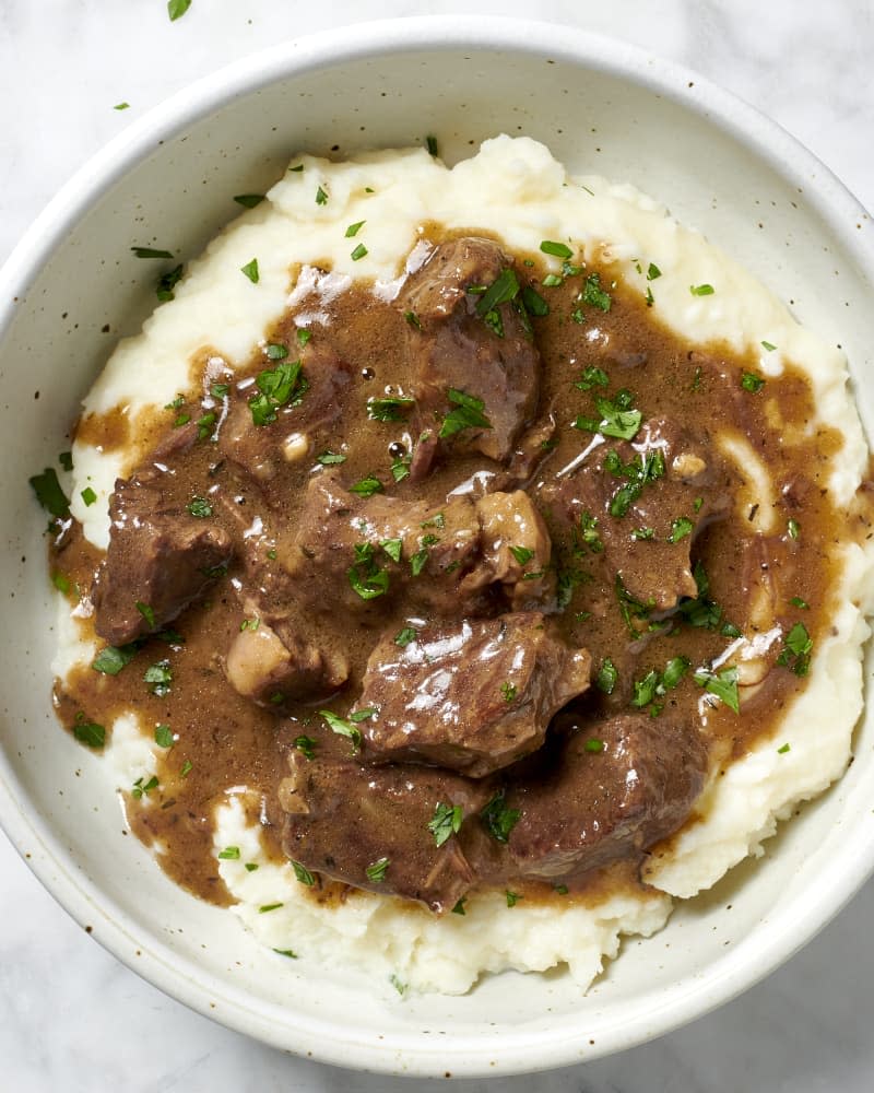 Overhead view of beef tips on mashed potatoes, topped with herbs in a white and brown speckled bowl.