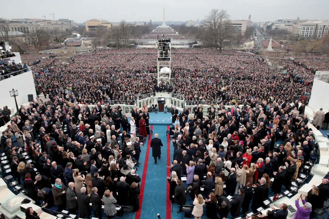 From above, Donald Trump on a blue carpet that leads to a podium surrounded by a crowd of people with the National Mall and Washington Monument visible in the background.