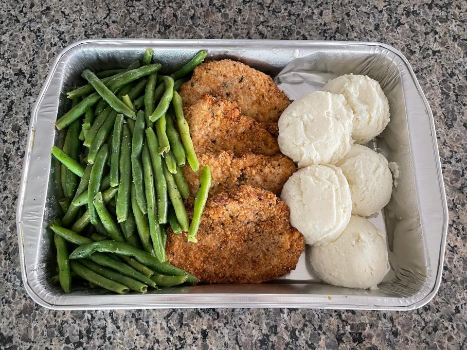 Four pieces of breaded chicken, scoops of mashed potatoes, and green beans in an aluminum container