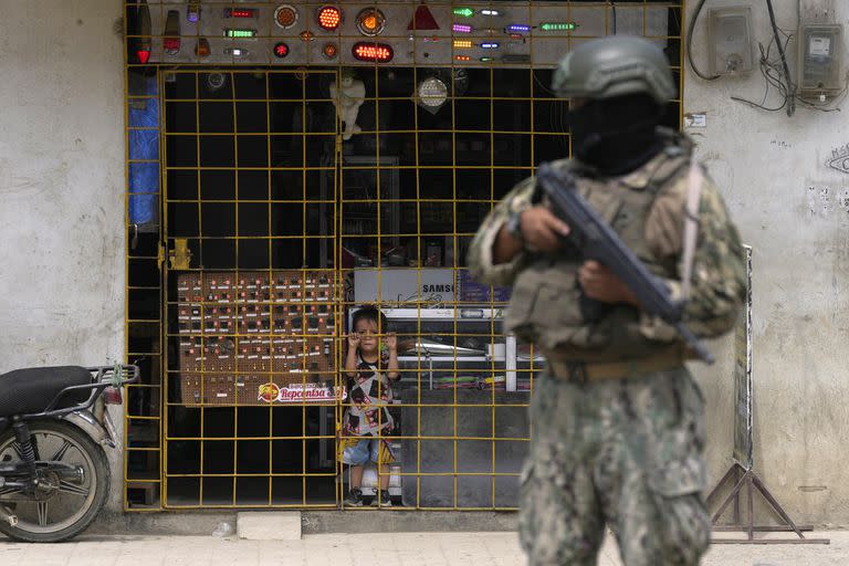 Un niño observa desde detrás del enrejado de una tienda mientras un soldado hace guardia en un puesto de seguridad, en Durán, Ecuador, el 14 de agosto de 2023. El presidente del país declaró el estado de excepción en algunas zonas luego del asesinato de un candidato presidencial tras un mitín en la campaña para los comicios adelantados, previstos para el 20 de agosto. (AP Foto/Martín Mejía)