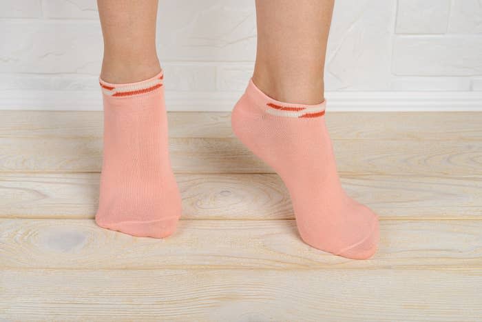 Pair of feet wearing ankle socks, both in a standing position, on a wooden floor. No people are identified in the image