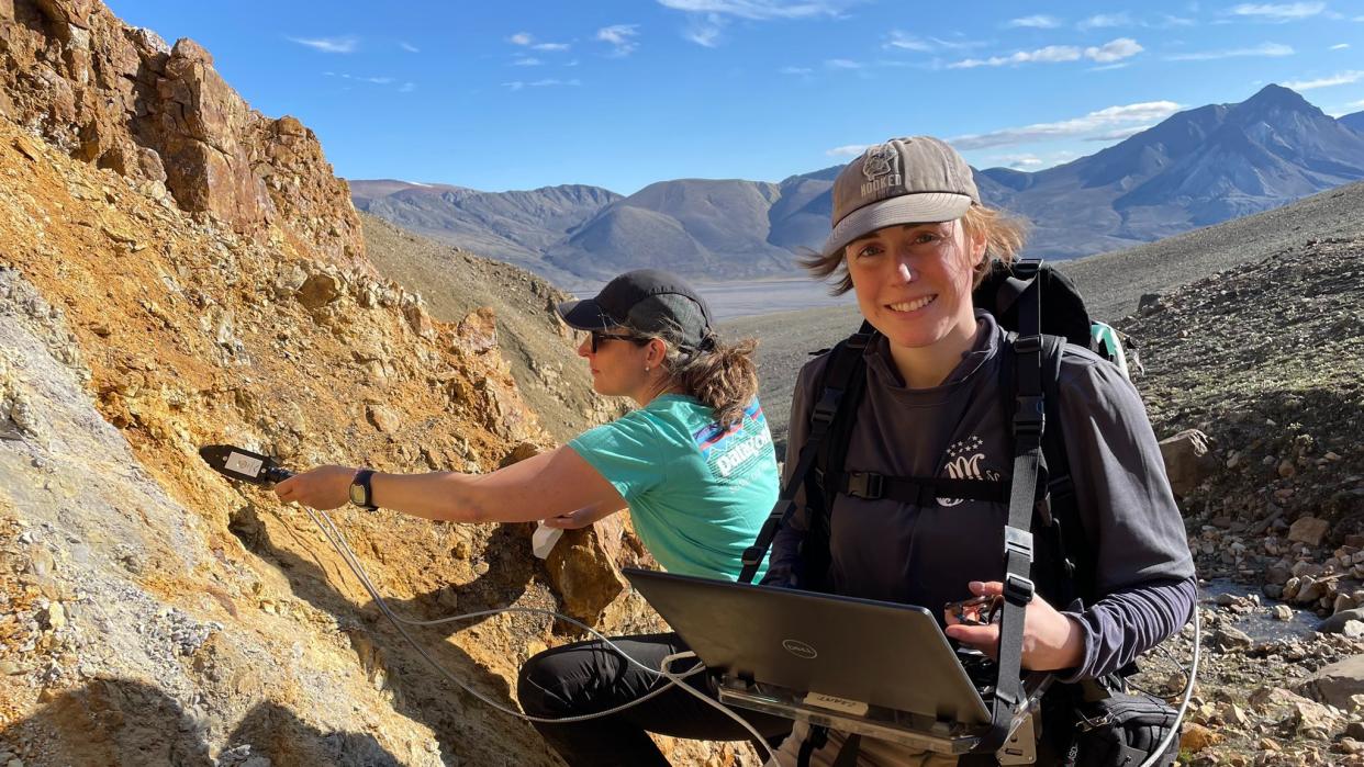  two women on a rocky outcrop. one squats in front with a laptop while the second, in behind, uses an instrument against a rusty slope. in the background are mountains and blue sky with clouds 