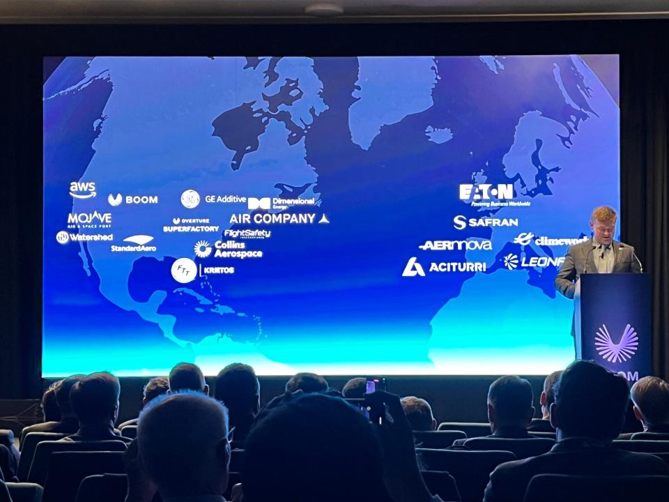 Boom Supersonic CEO Blake Scholl speaks at a podium at the Paris Air Show, while a map of its partners is displayed