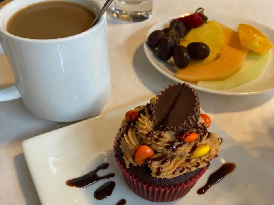 A chocolate cupcake topped with a light brown frosting with orange and yellow M&Ms, a mini Reese's Pieces, and a chocolate drizzle. It's surrounded by a cup of coffee and a plate of grapes, strawberries, pineapple, cantaloupe, melon, and an orange.