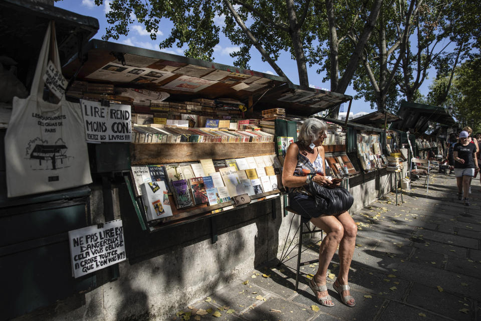 A woman rests along bookseller booths, called "bouquinistes" at the Seine Riverbank in Paris, Tuesday, Aug. 22, 2023. The host city of Paris vowed to deliver an extraordinary grand opening on July 26, 2024, as the ceremony is expected to draw about 600,000 spectators to the Parisian quayside. Citing security measures, the Paris police prefecture ordered on July 25 the removal of 570 stationary boxes out of which booksellers have operated for decades on the quays of the Seine river. (AP Photo/Sophie Garcia)