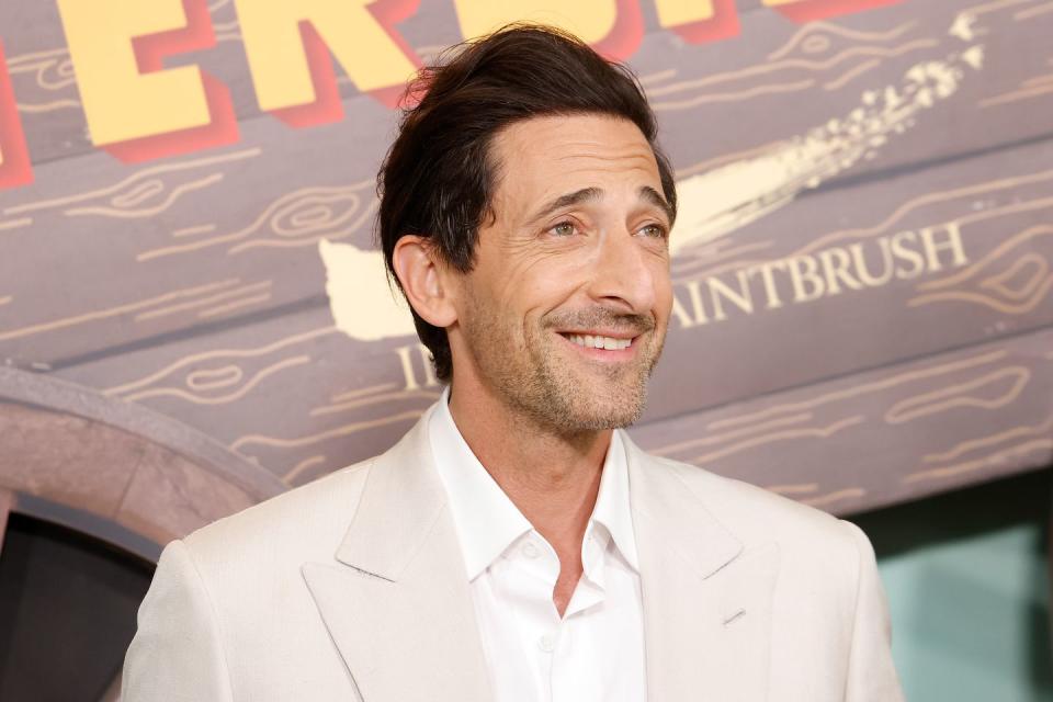 adrien brody smiles and wears a cream colored suit jacket with a white collared shirt