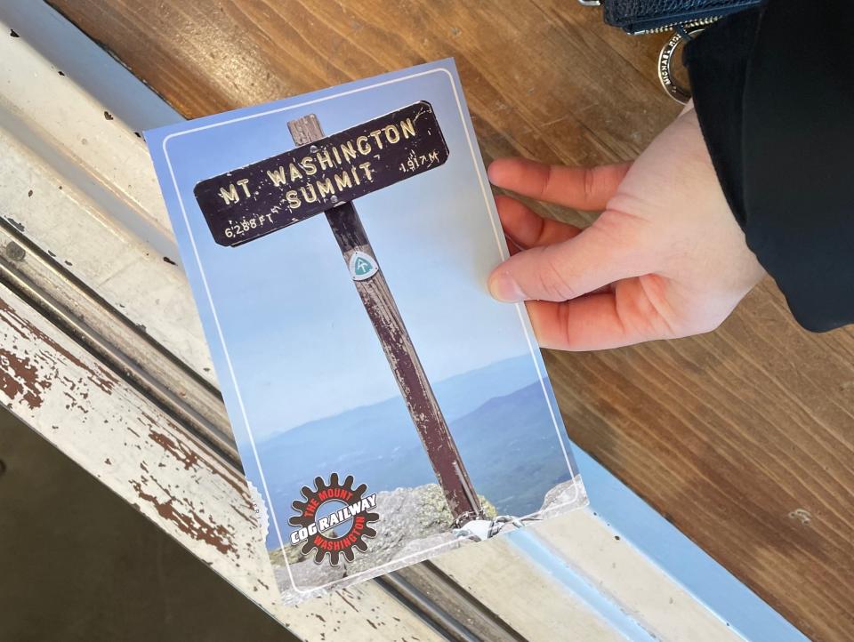 Hand holding a postcard of a sign reading "Mt. Washington Summit."