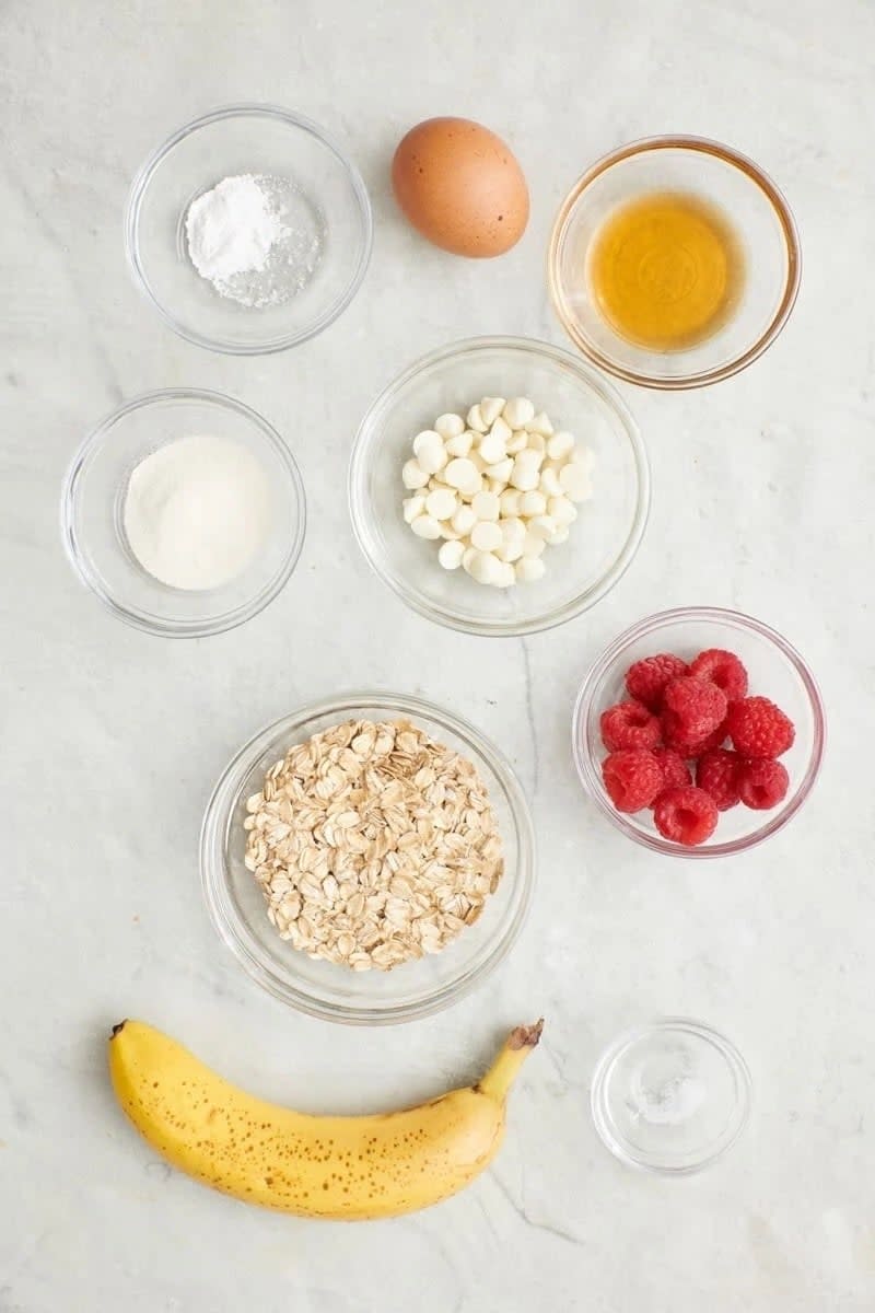 Oatmeal, raspberries, a banana, egg, white chocolate chips, baking powder, vanilla extract, and powdered ingredients in bowls arranged on a marble surface