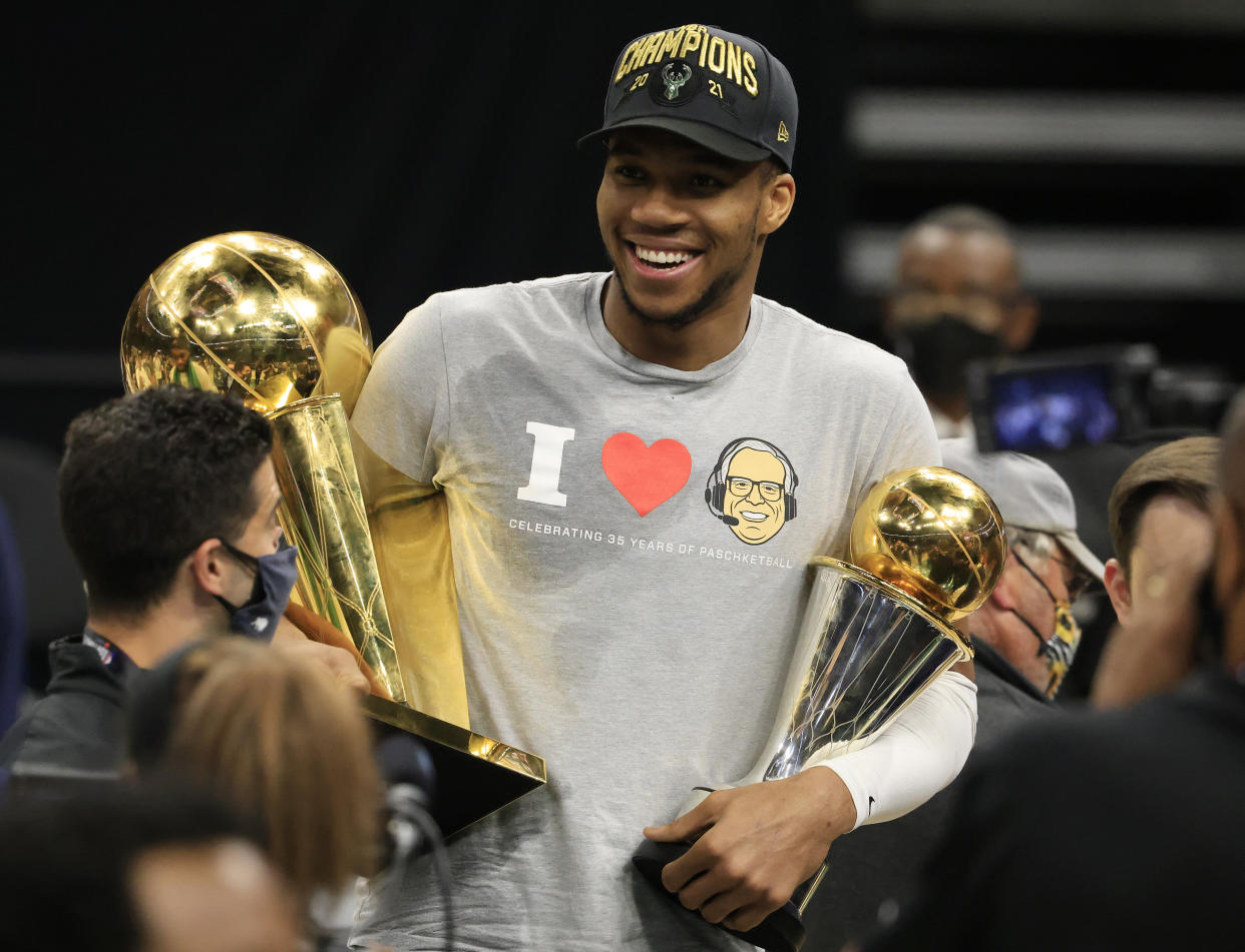 Giannis Antetokounmpo smiles as he holds the Bill Russell NBA Finals MVP Award and the Larry O'Brien Championship Trophy.