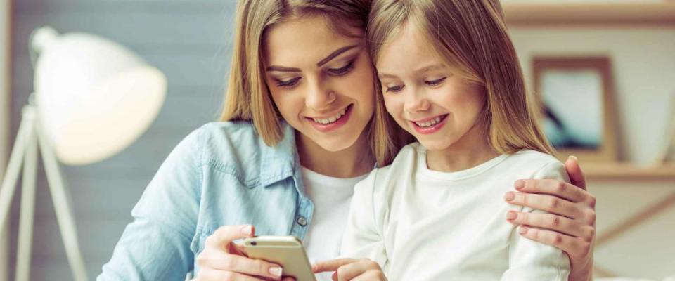 Beautiful young mom and her little daughter are using a smartphone and smiling while sitting on sofa at home