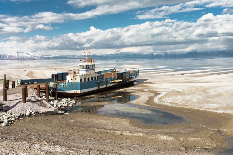 Rusting boat on salty ground, lake and mountains in distance.