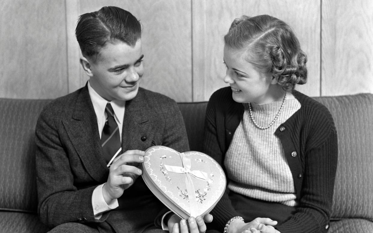 A teenage boy gives a girl a heart-shaped box of chocolates in the 1930s - Getty Images Fee