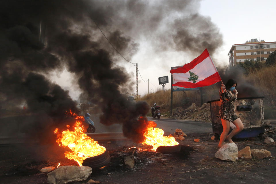 An anti-government protester carries her national flag past burning tires blocking the main highway during protests against the corruption in Khaldeh, south of Beirut, Lebanon, Wednesday, Nov. 13, 2019. A local official for a government political party was shot dead by soldiers trying to open a road closed by protesters in southern Beirut late Tuesday, the army reported, marking the first death in 27 days of nationwide protests. (AP Photo/Hussein Malla)