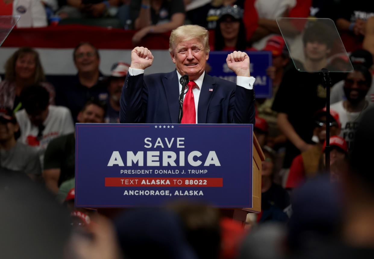 Donald Trump raises both fists in the air in an expression of triumph at a podium marked: Save America, President Donald J. Trump, Text Alaska to 88022, Anchorage, Alaska.
