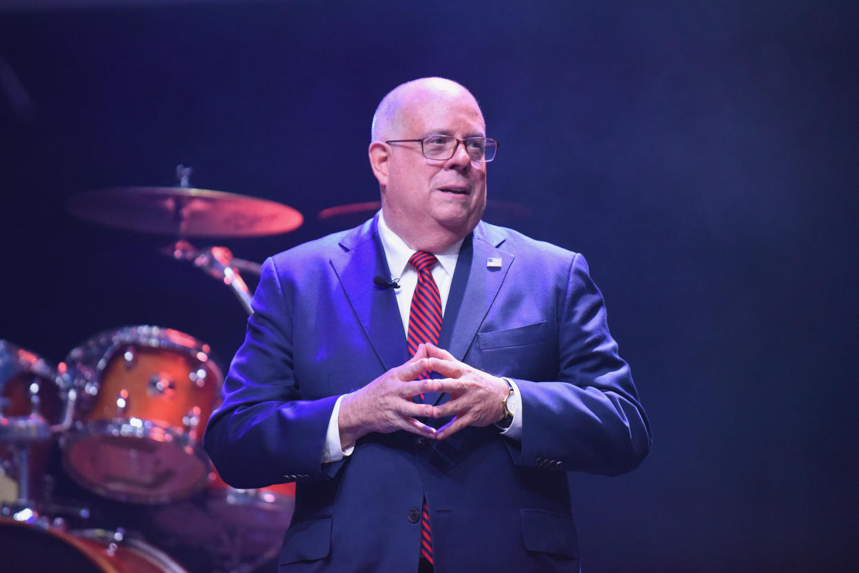 Gov. Larry Hogan makes a point, his fingertips pressed together, with a drum set behind him.
