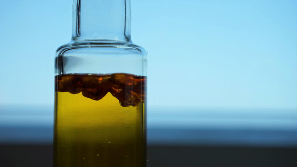 Glass bottle containing olive oil with floating herbs or spices, set against a blurred background