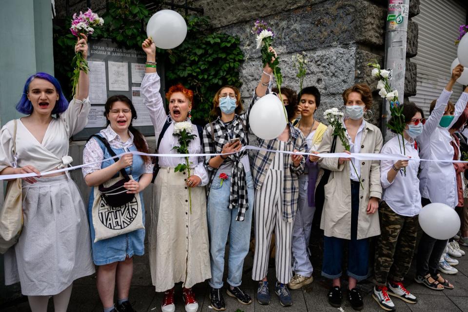 Women dressed in white stand in a line and shout while carrying flowers and balloons