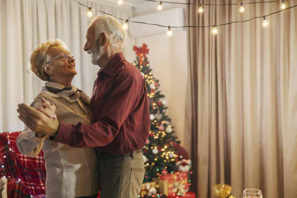 An elderly couple dance joyfully in a warmly decorated room with a Christmas tree and string lights in the background