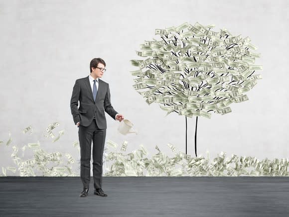 A businessman waters a tree with leaves made of dollar bills.