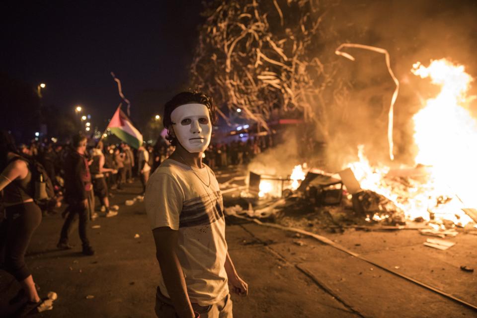 A masked anti-government protester stands by a burning barricade in Santiago, Chile, Monday, Oct. 28, 2019. Fresh protests and attacks on businesses erupted in Chile Monday despite President Sebastián Piñera's replacement of eight important Cabinet ministers with more centrist figures, and his attempts to assure the country that he had heard calls for greater equality and improved social services. (AP Photo/Rodrigo Abd)