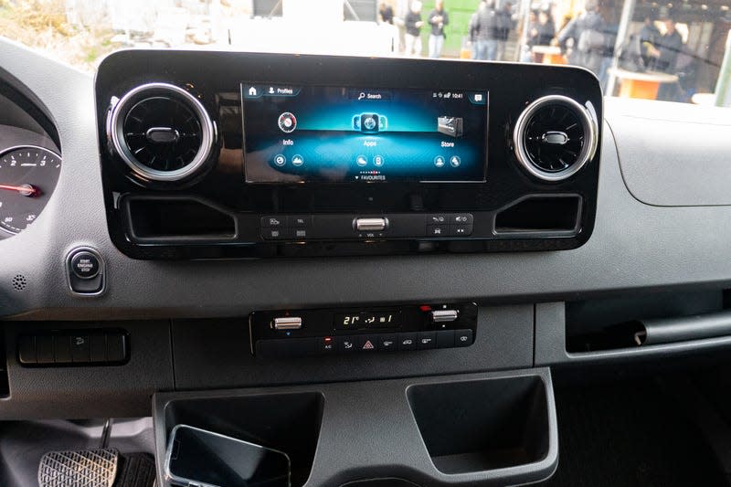 a close-up shot of the dashboard of a 2023 mercedes benz sprinter awd van, focusing on the rectangular landscape-oriented touchscreen in the middle of the dash. the screen is showing audio info, and is flanked on either side by circular air-conditioning vents