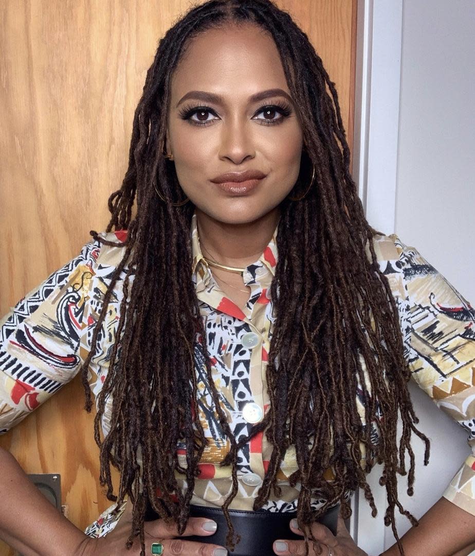 photo of filmmaker Ava DuVernay, a Black woman with long locs standng in front of a door. She is wearing a colorful printed shirt. 