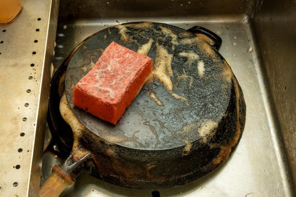 A dirty cast iron skillet upside-down, with suds and a pink sponge on top.