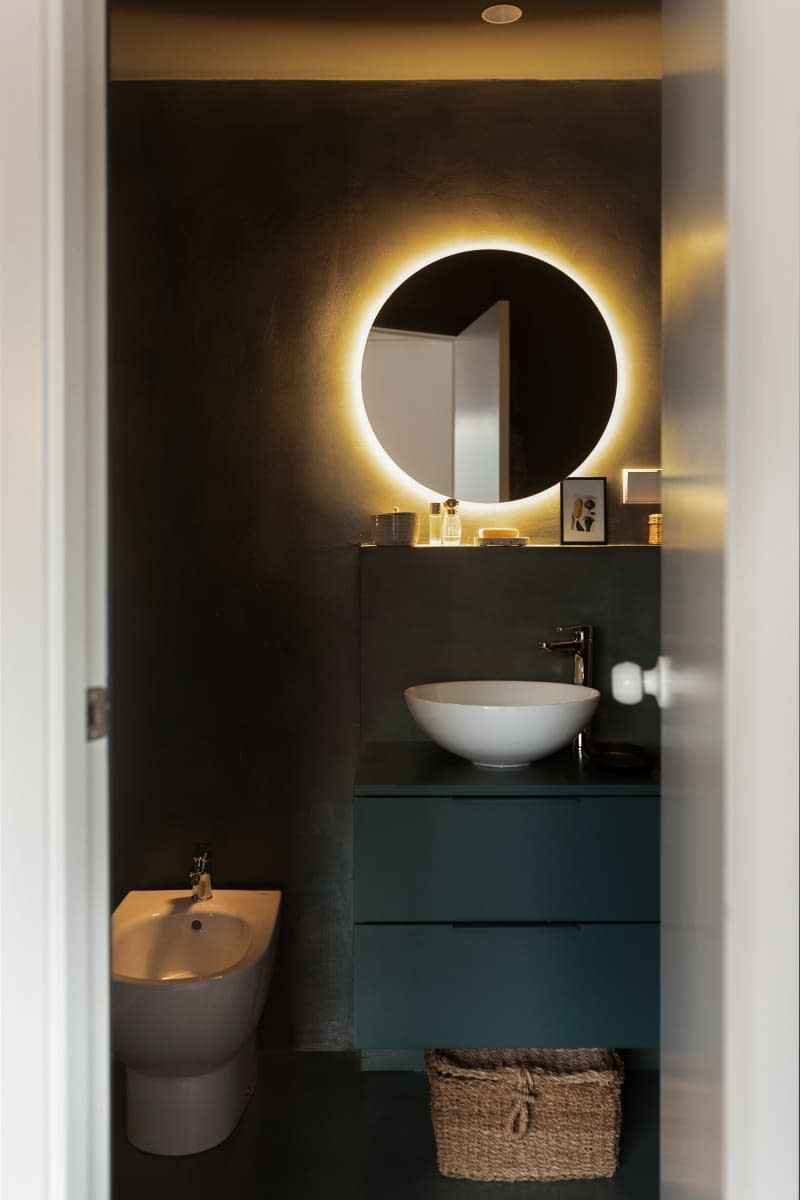 A black bathroom with circular mirror with light and white fixtures.