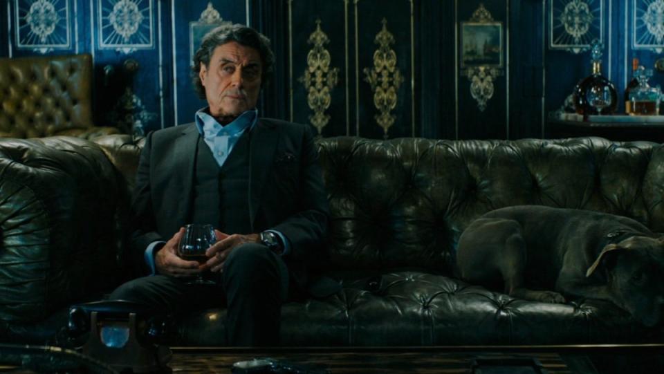 Ian McShane as Winston sits on a couch with a drink in the Continental in John Iwck: Chapter 3