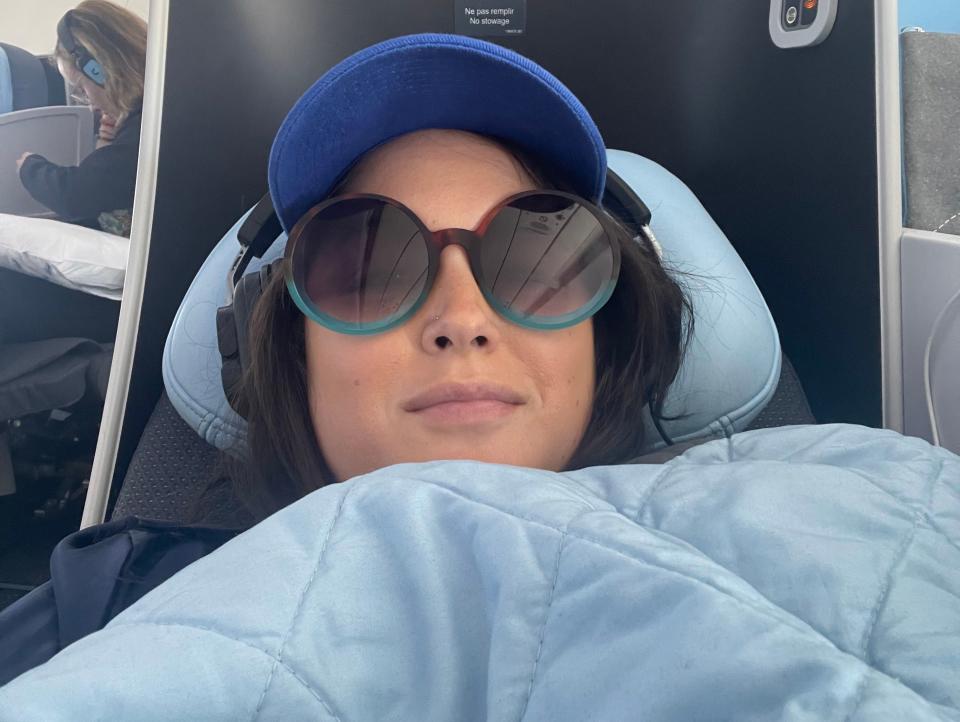 Katka, wearing sunglasses and chunky headphones, relaxes under a blue blanket in a business-class seat.