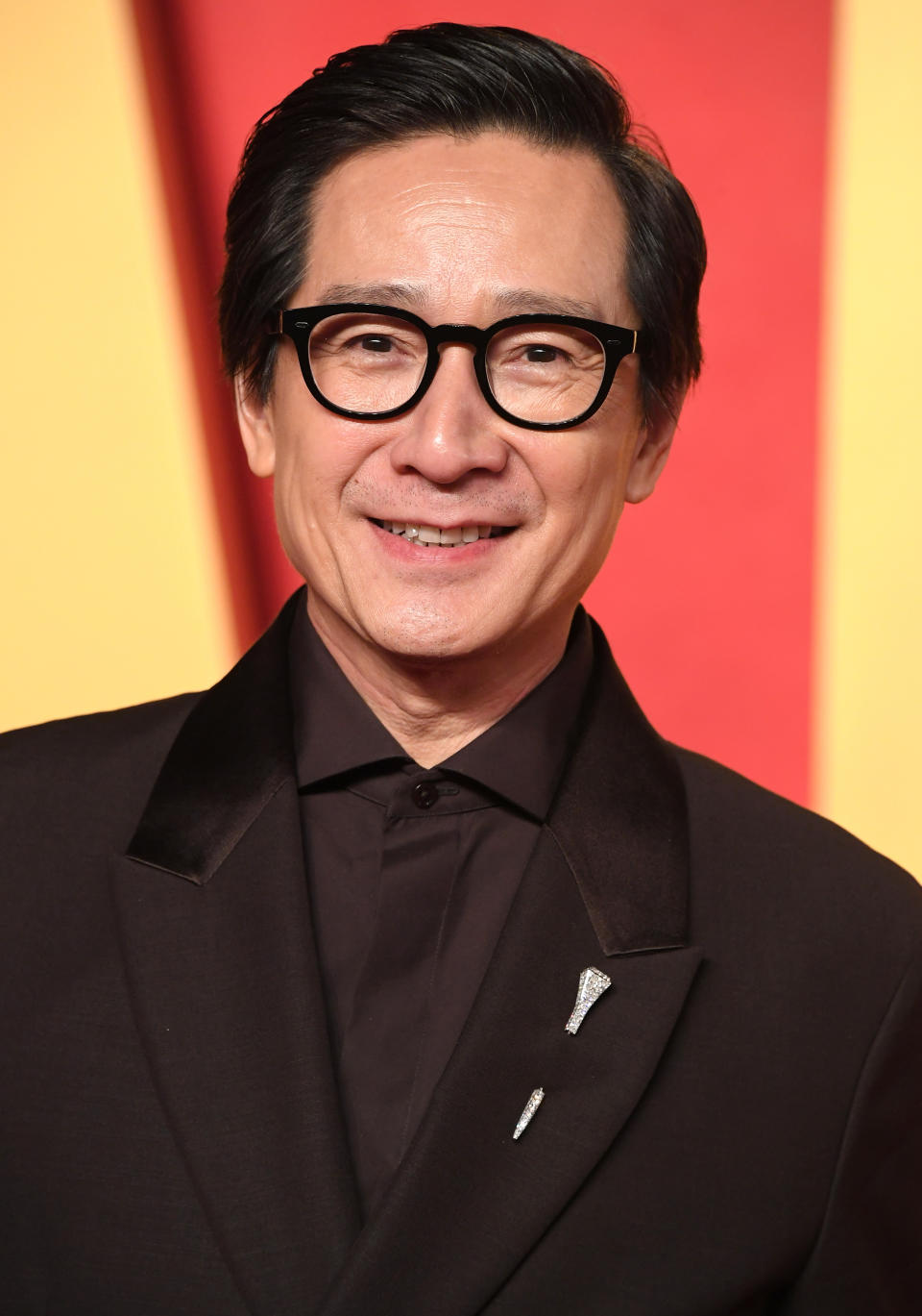 Person wearing glasses and a dark suit with a black satin lapel and a decorative pin, smiling on a patterned background