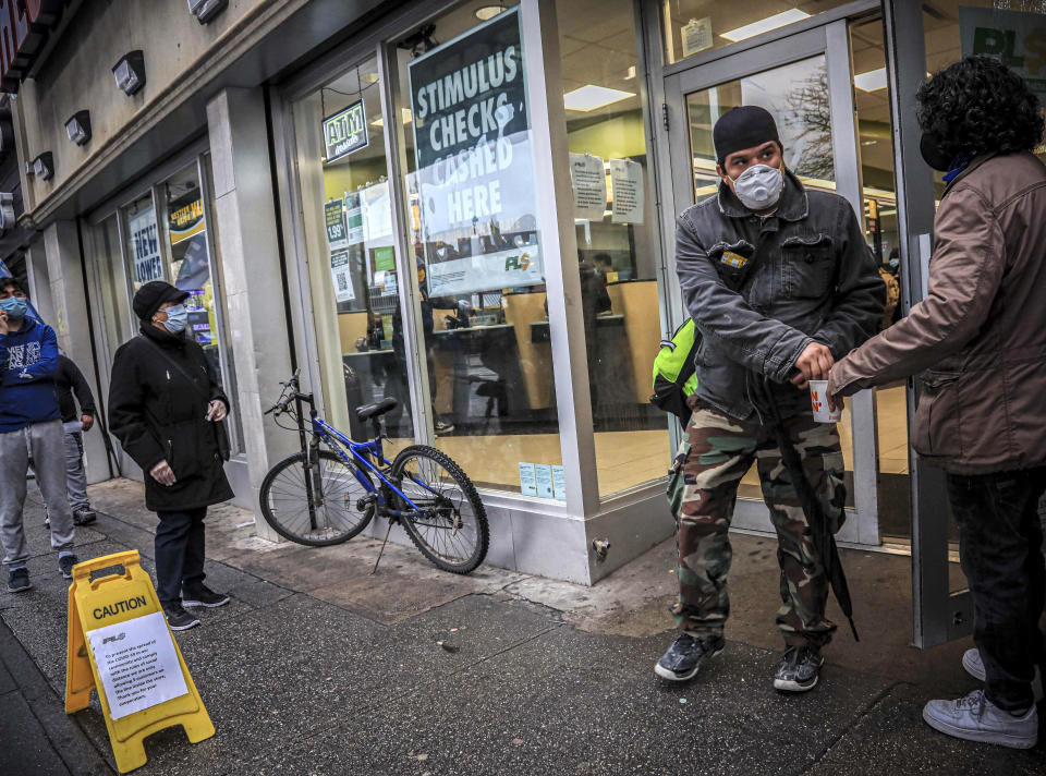 In this April 24, 2020, photo, a man holds out a cup for money donations, left, as customers wearing face mask due to COVID-19 concerns, exit a check cashing service center in the Brooklyn borough of New York. While millions of U.S. workers have already received a quick relief payment from the federal treasury through direct deposit, millions of others without traditional bank accounts must wait weeks for paper checks. Advocates for the poor say this is an opportunity to get many of those who are unbanked into the formal financial system. (AP Photo/Bebeto Matthews)