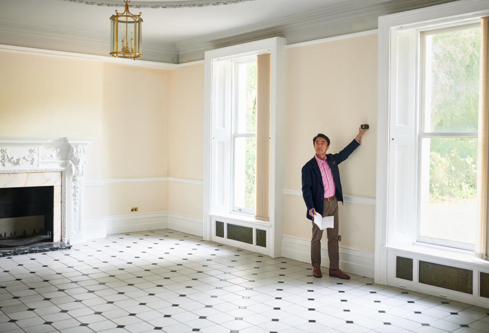 A person stands in an empty room with a fireplace and large windows, adjusting a thermostat on the wall. They hold a piece of paper in the other hand