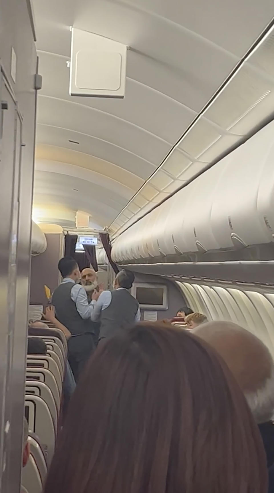 In this image from a video, a passenger, center, speaks with crew members during a flight from Australia to Malaysia, Monday, Aug. 14, 2023. Malaysia Airlines flight MH122 returned on Monday to Sydney, where the passenger was arrested hours later in what police described as an emergency incident. (Velutha Parambath via AP)