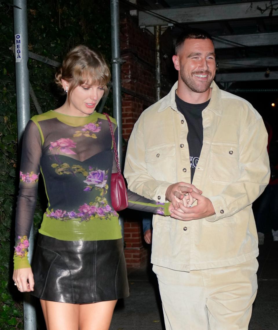 Taylor Swift in a floral mesh top and leather skirt holds hands with Travis Kelce in a beige jacket and trousers