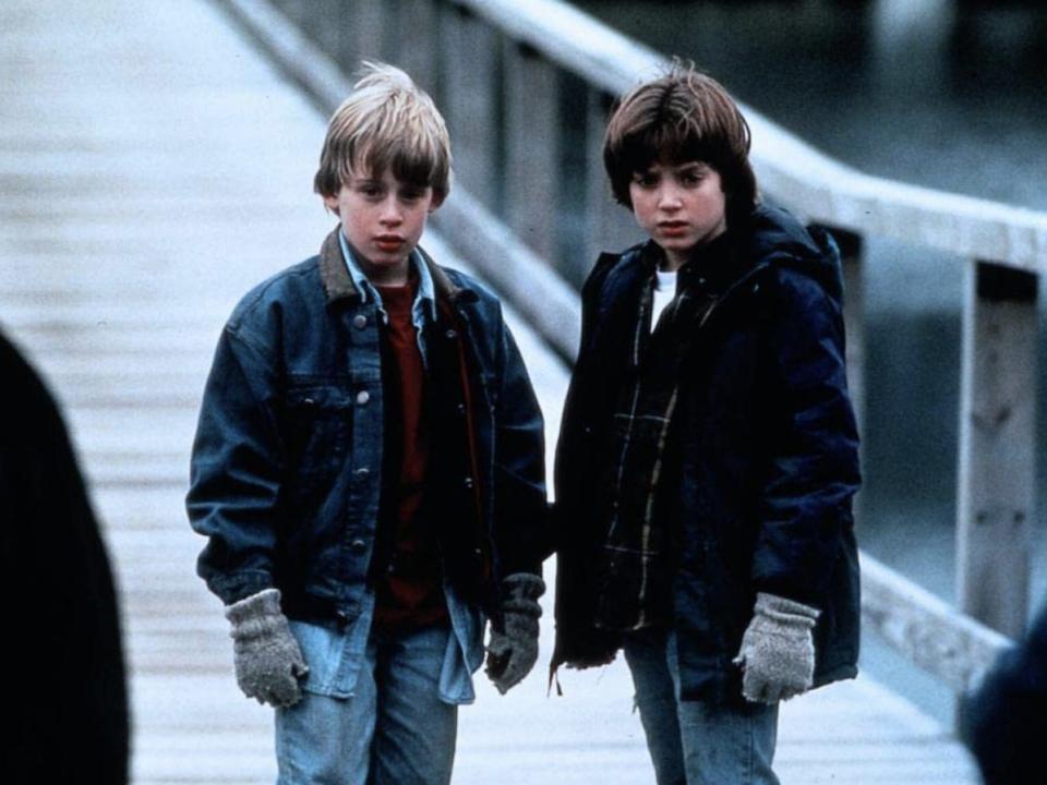 A young Macaulay and Elija in coats, jeans, and gloves looking in the same direction on a pier.