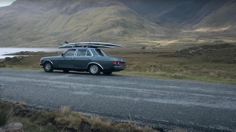 A restored 1980s Mercedes sedan with surfboards on the roof is parked in a mountainous area near water